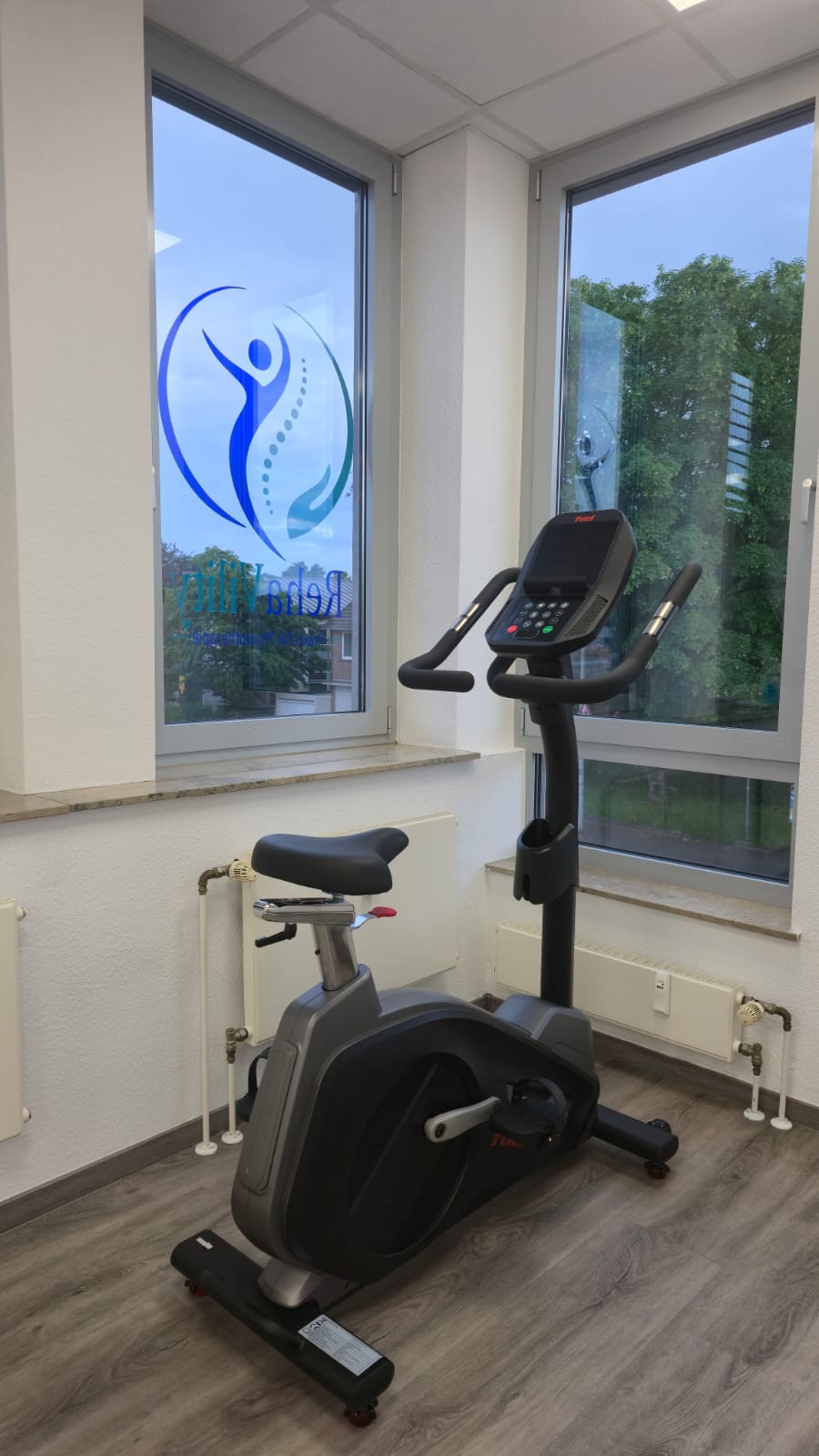Trainingsbereich mit Ergometer und Praxislogo am Fenster.